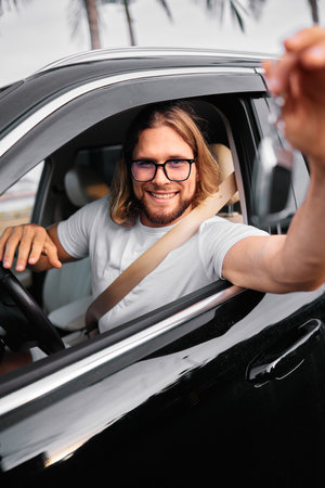 Happy man in casual attire sitting in a car, smiling and holding car keys Tropical scenery in the...