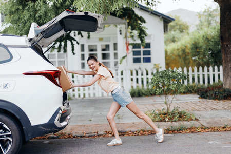 Delivery, woman unloading, car boot, residential background, sunny day A young woman in casual wear is pulling a box from a car trunk amidst greenery and a house in the backgroundの写真素材