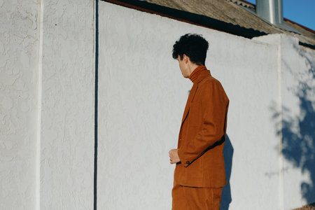 A stylish young man in a brown suit standing against a textured white wall, showcasing modern...
