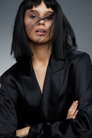 Blackhaired woman in a black suit poses confidently with arms crossed against a gray background, conveying professional strength and determinationの写真素材