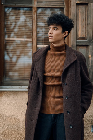 Fashionable young man in brown coat and black turtleneck sweater standing in front of window with old wooden doorの写真素材