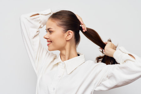 Young woman smiling while tying her long brown hair in a ponytail against a light gray background, showcasing natural beauty and confidence Portrait shot with focus on hairstyleの写真素材