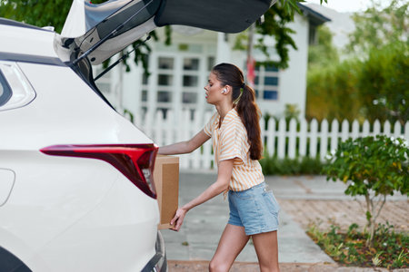 Young woman loading box into car trunk, outdoor setting, casual attire, summer day Bright and sunny, green trees in the background Focused on the task at handの写真素材