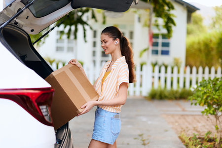 Young woman loading a cardboard box into her car, working outdoors She wears casual summer attire with a cheerful expression, surrounded by greenery and a cozy home backdropの写真素材
