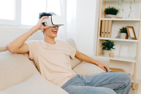 Young man enjoying virtual reality experience at home, sitting on a cozy sofa in a well lit, modern...