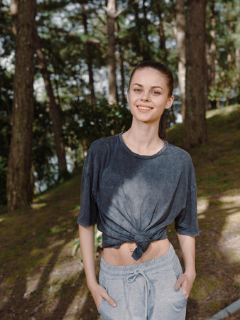 Smiling young woman in casual outfit, standing outdoors in the forest, enjoying nature and a...