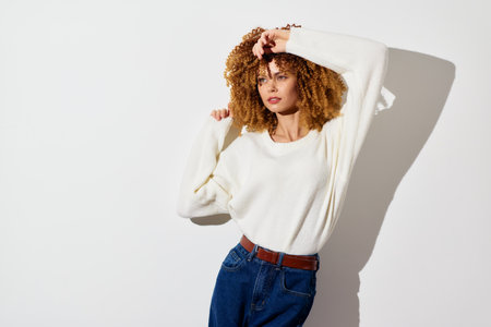 Fashionable young woman with curly hair in a cozy white sweater posing against a plain light...