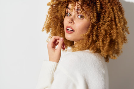Stylish young woman with curly hair wearing a white sweater, against a neutral background,...