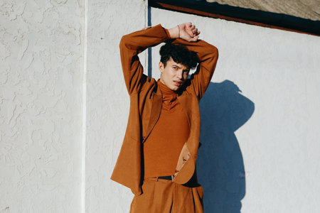 Young man posing with arms raised against a textured white wall He wears a stylish outfit in warm earthy tones, creating a modern and artistic vibeの写真素材