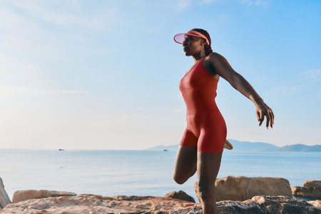 Woman in red athletic outfit practicing yoga on rocky shore at sunriseの写真素材