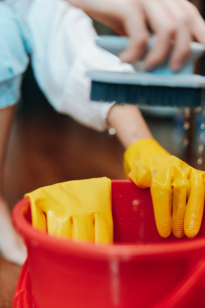 Person in yellow rubber gloves cleaning a red bucket with a brush on wooden floor in front of another person in white shirt and blue jeansの写真素材