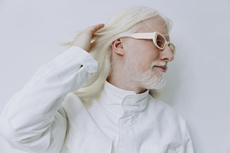 Stylish elderly man with long white hair and glasses poses thoughtfully against a plain white...