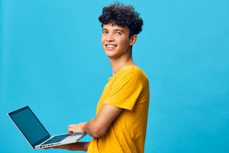 Young man with laptop smiling against a blue background, casual wear, cheerful expression,...