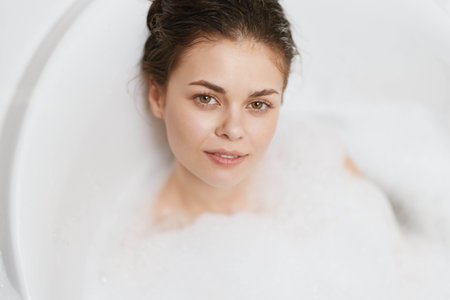 Relaxed young woman enjoying a bubble bath, serene expression on her face, surrounded by white foam....