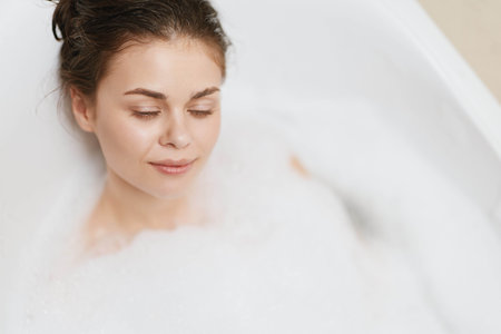 Relaxed young woman enjoying a bubble bath at home, eyes closed with a serene expression, creating a...