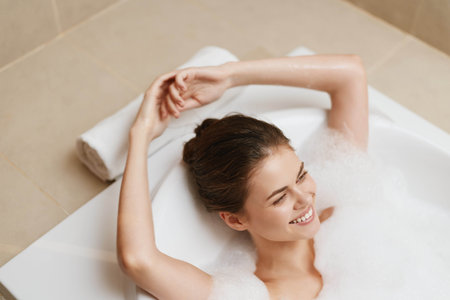 Relaxed young woman enjoying a bubble bath at home, smiling, surrounded by soft towels and warm...