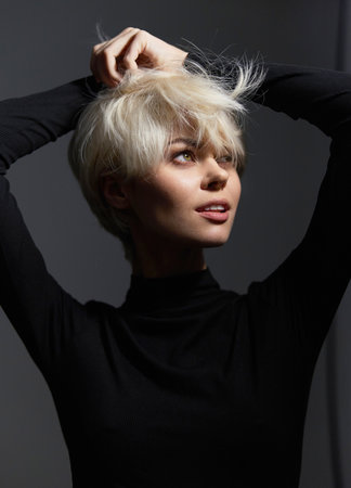 Confident young woman with short blonde hair playfully adjusting her hair while wearing a black turtleneck. Studio portrait with soft lighting and neutral background.の写真素材