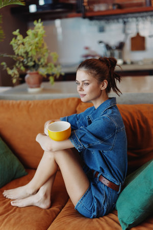 Woman sitting on couch with coffee cup in hand, plant in background cozy home interior sceneの写真素材