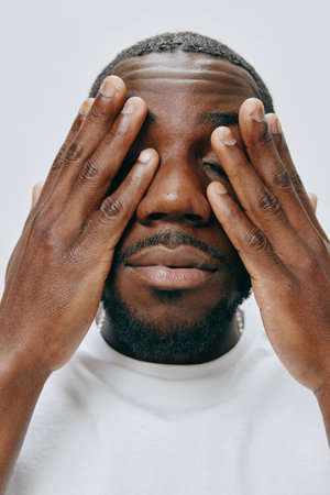 portrait of a young man covering his eyes, expressing emotions like surprise or thoughtfulness on a neutral background, wearing a casual white t shirt, showcasing a heartfelt momentの写真素材