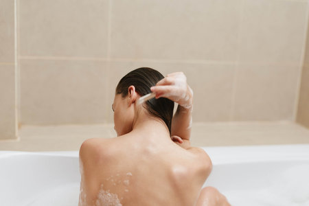 Relaxed young woman with wet hair applying shampoo while enjoying a bath, creating a calming...
