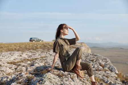 Woman sitting on rock with hands on head and car in background reflecting on travel and adventureの写真素材