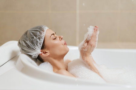Relaxed woman enjoying bubble bath in modern bathroom, wearing shower cap and holding bubbles,...