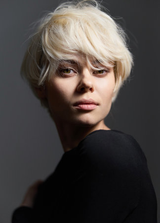 Stylish young woman with short blonde hair gazing over her shoulder, showcasing elegance and confidence against a dark, moody backdrop. Portrait and beauty concept.の写真素材