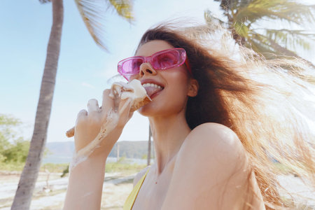 Woman enjoying ice cream on a sunny beach with palm trees in the background, wearing stylish...