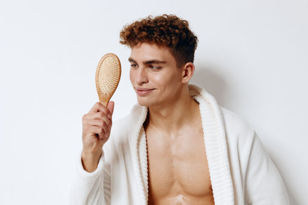Confident young man examining wooden hairbrush while wearing a cozy bathrobe, suggesting relaxation and self-care at home. Personal grooming concept.の写真素材