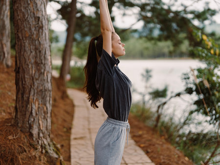 Young woman practicing yoga outdoors by the lake, stretching arms up to the sky, enjoying nature and tranquility Wellness and mindfulness conceptの写真素材