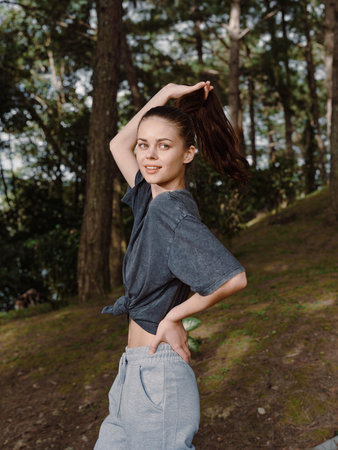 Young woman with long hair casually tying it back while standing in a forest, wearing a relaxed...