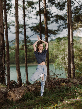 Young woman practicing yoga in nature, balancing on one leg with hands raised, surrounded by trees near a calm lake Wellness and mindfulness conceptの写真素材