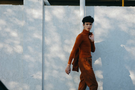 Stylish young man walking near a textured white wall, wearing a rust colored outfit, showcasing modern fashion and confidence in an urban settingの写真素材