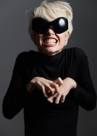 Playful European woman with short blonde hair wearing oversized black sunglasses and a black turtleneck sweater, expressing a fun and quirky emotion against a dark background.の写真素材