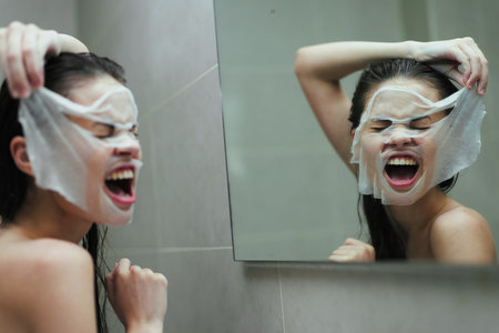 Excited Asian woman having fun while applying a skincare mask in the bathroom, looking in the mirror. Self-care and beauty routine concept.の写真素材