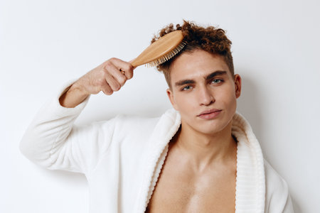 Stylish young man with curly hair brushing his hair in a cozy bathrobe, enhancing his grooming routine. Personal care and self-confidence concept.の写真素材