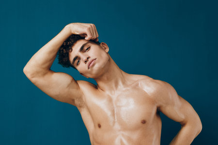 Confident young man with curly hair posing shirtless against a teal background, showcasing a strong physique and a relaxed expression. Fitness and self-esteem concept.の写真素材