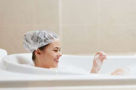 Smiling young woman enjoying a relaxing bath, wearing a shower cap and playfully splashing water with her hands. Self-care and relaxation concept.の写真素材