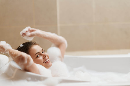 Joyful young woman relaxing in a bubble bath, creating a serene atmosphere with foam surrounding her. Self-care and wellness concept at home.の写真素材