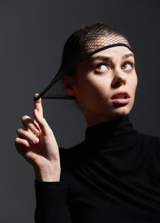 Pensive young woman in black turtleneck and fishnet headpiece, looking upward with an expression of curiosity. Fashion and emotion concept in a dark setting.の写真素材
