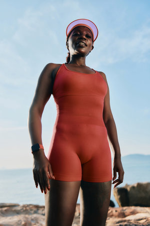Athletic woman in a sporty outfit standing by the water with a confident pose against a clear blue...