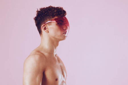 Stylish young man with curly hair wearing bold red visor sunglasses, showcasing a modern fashion statement against a soft pink background.の写真素材