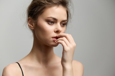 Thoughtful young woman with a neutral expression, biting her finger gently. Close-up portrait with soft lighting on a light gray background, capturing vulnerability and introspection.の写真素材