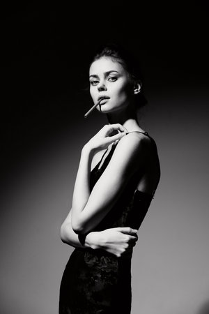 Elegant woman in a sleek black dress posing confidently with a cigarette, showcasing a bold and rebellious attitude. Dramatic lighting enhances her striking features.の写真素材