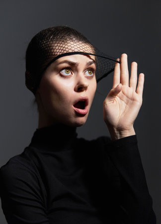 Surprised woman in black turtleneck and fishnet veil, expressing shock with hand raised. Dramatic lighting adds depth to the emotional portrait.の写真素材