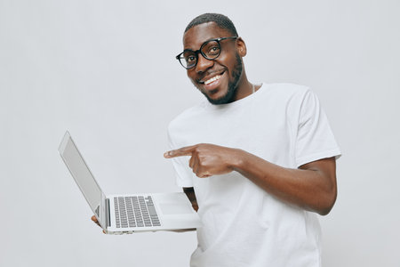smiling man pointing at laptop, casual wear, white background, positive emotions, technology engagement, African descent model, modern lifestyle, professional setting, cheerful expression, connectionの写真素材