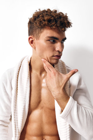 Thoughtful young man with curly hair in a white robe, gazing thoughtfully while touching his chin. Self-care and reflection concept.の写真素材