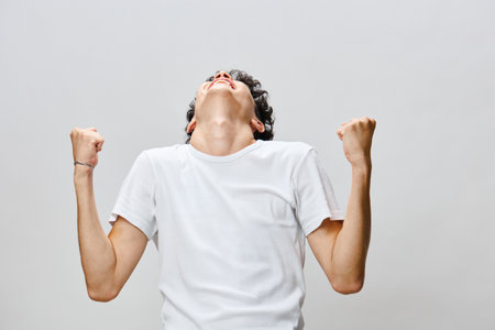 Excited young man celebrating with raised fists, showing joy and enthusiasm against a plain light background, conveying victory and positive emotionsの写真素材