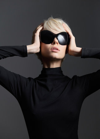 Confident woman wearing stylish black sunglasses and a black turtleneck, striking a bold pose against a dark gray background. Fashion and elegance concept.の写真素材