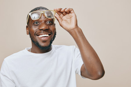 Happy African man with glasses, joyful expression, wearing casual white t shirt, beige background, showcasing excitement and positivity in a friendly atmosphereの写真素材